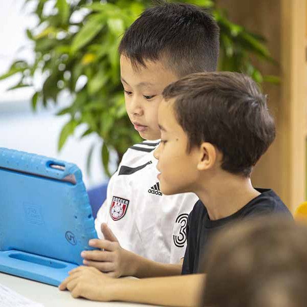 Two young boys are sitting at a table looking at a tablet computer.