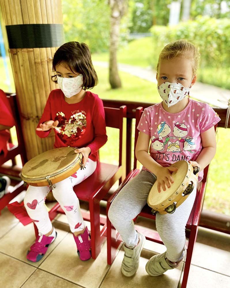 Two little girls wearing face masks are sitting on chairs playing tambourine.