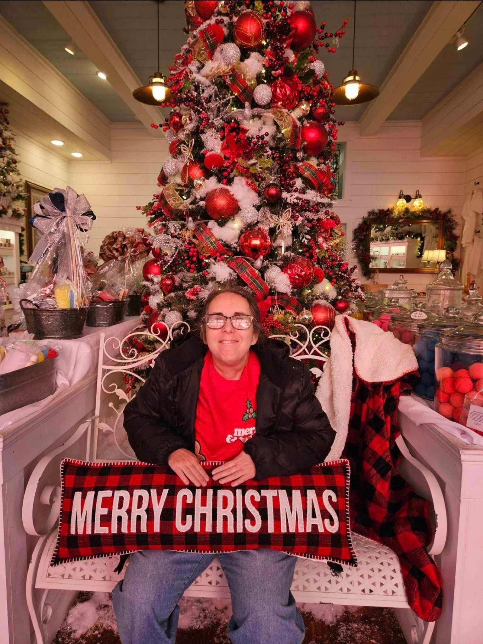 a woman is sitting in front of a christmas tree holding a merry christmas sign .