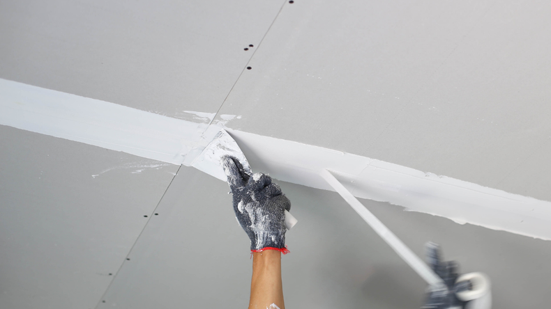 A person wearing gloves applies joint compound with a taping knife along a seam between grey drywall ceiling panels.