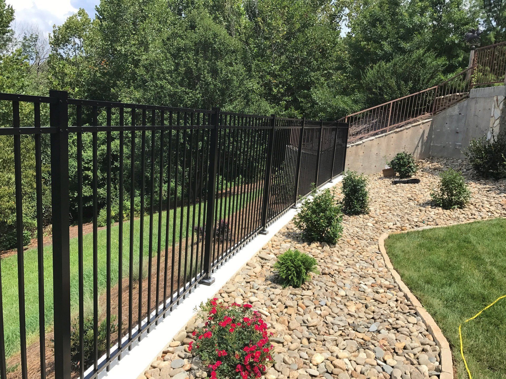 Black metal fence along a landscaped yard with rock and bushes; trees in the background.