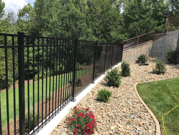 Black metal fence along a landscaped yard with rock and bushes; trees in the background.