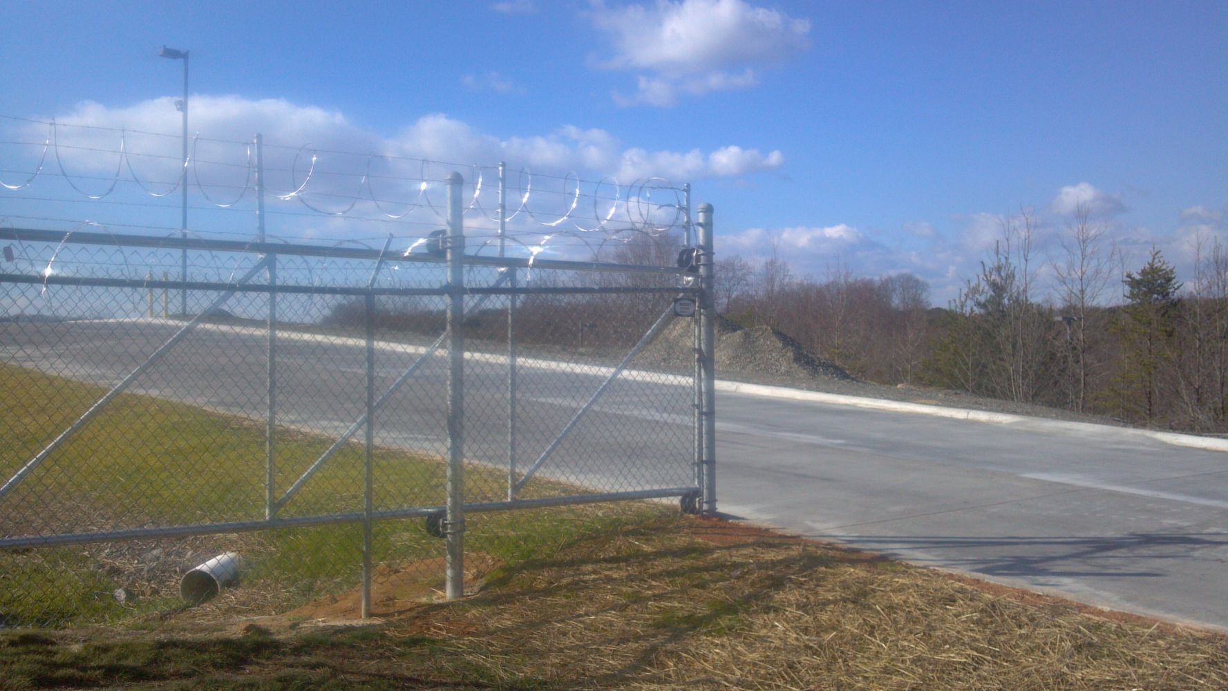 Chain link fence topped with barbed wire, bordering a paved road under a blue sky.