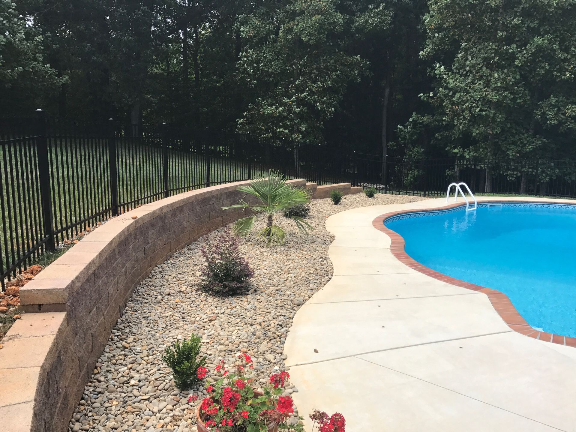 Poolside landscaping: a curved retaining wall, fence, and pool with blue water, trees, and flowers.