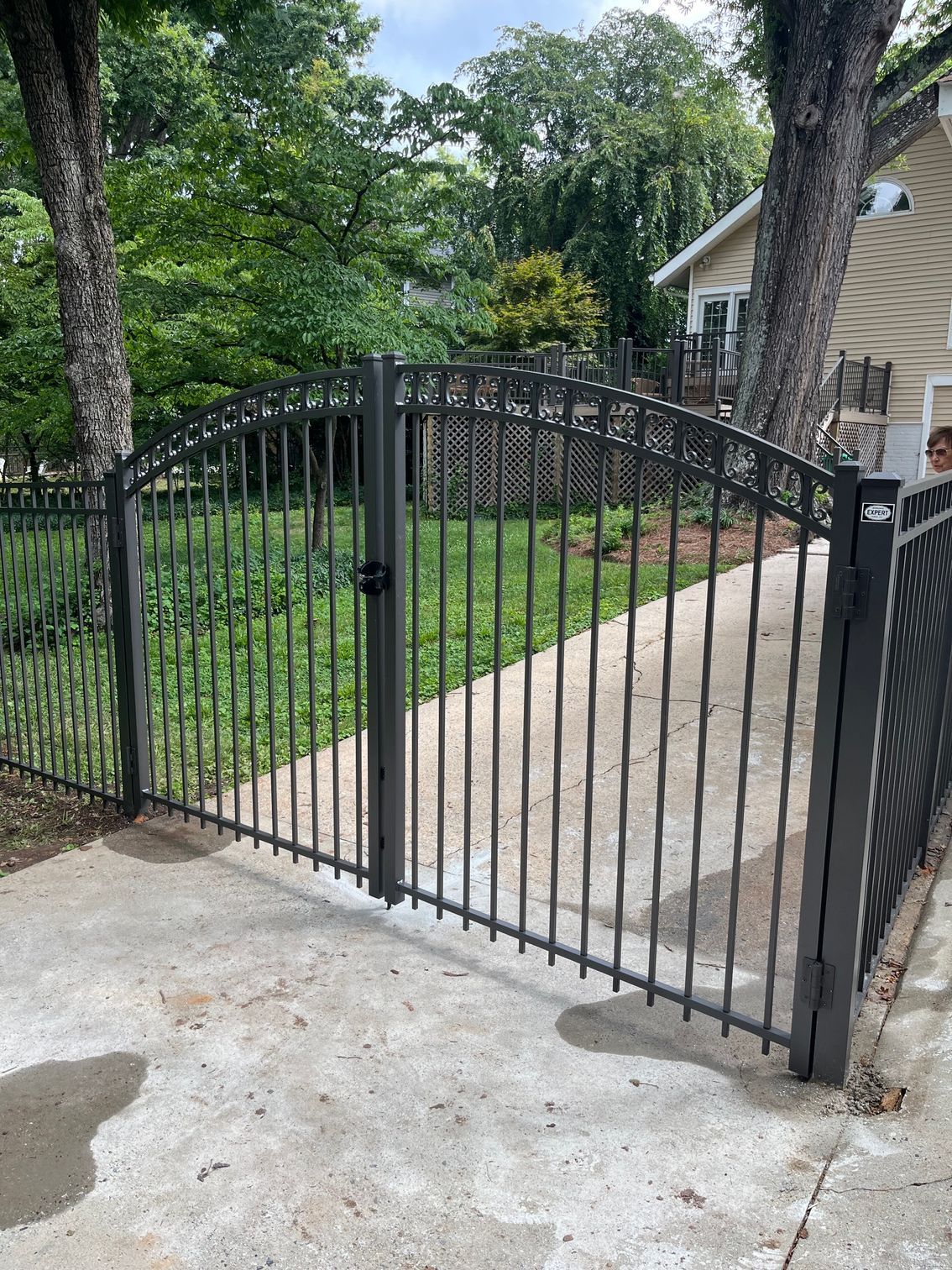 Black metal gate leading to a grassy yard. Two trees flank the sides and a beige house is in the background.
