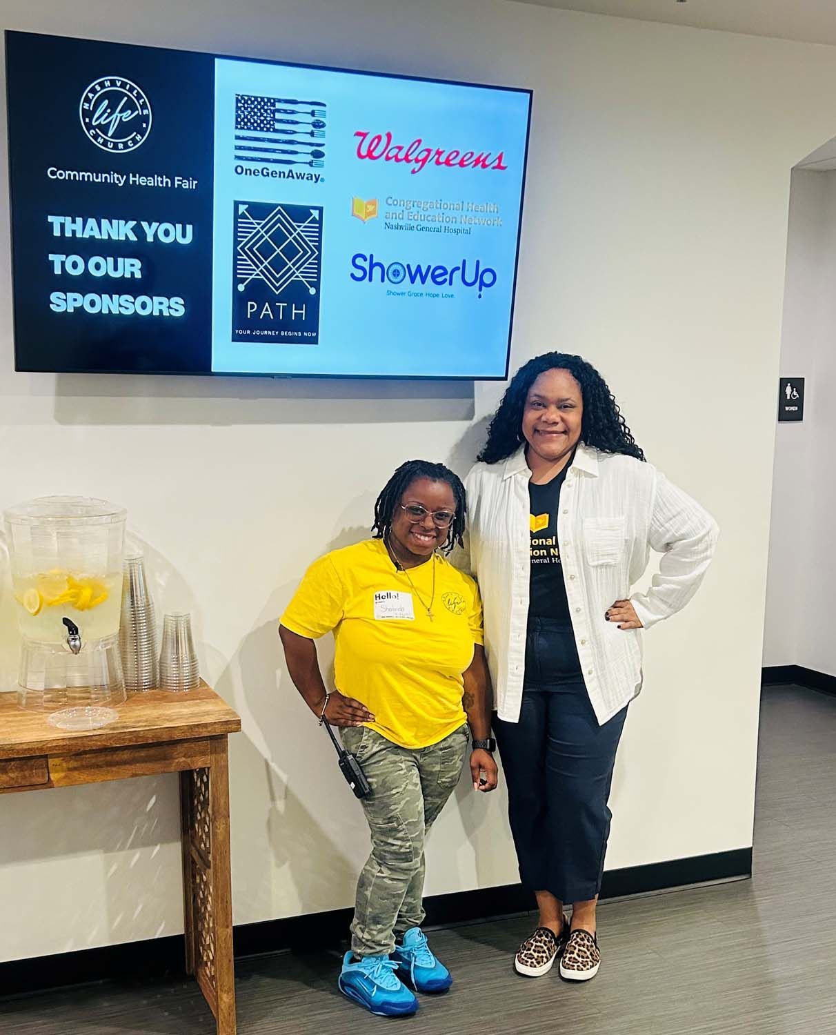 Two people stand smiling in front of a monitor displaying sponsor logos for a Community Life event.
