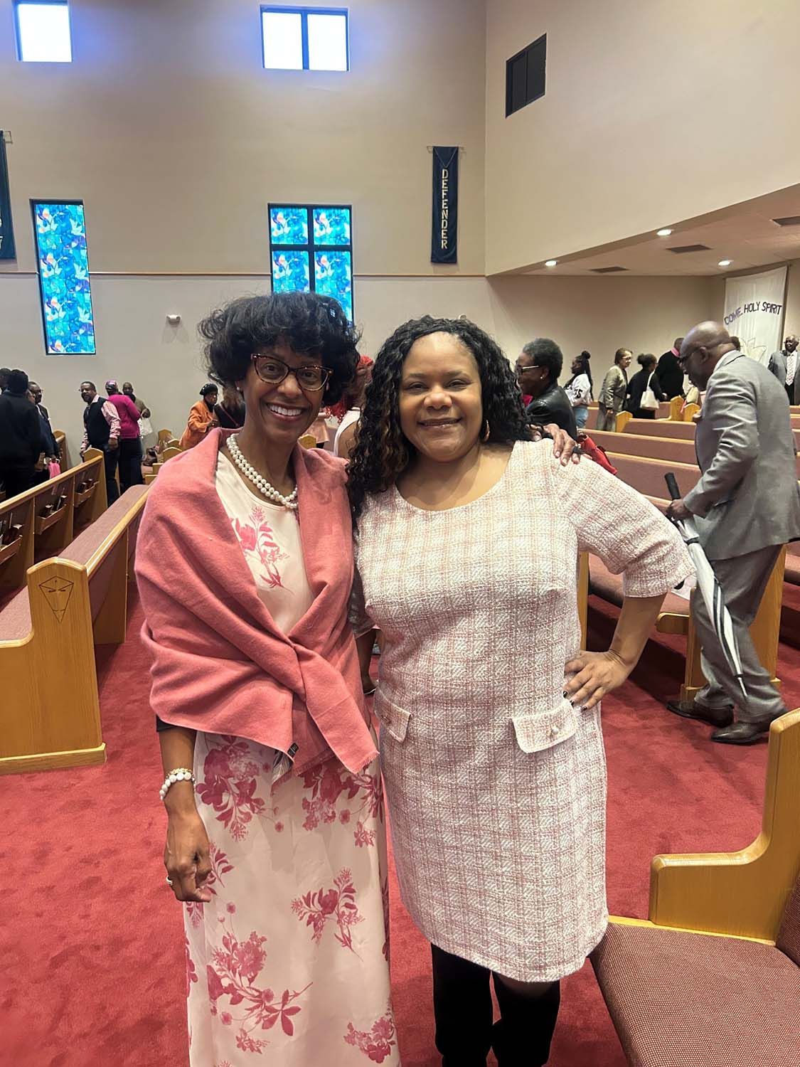 Two smiling individuals stand in a church aisle, wearing dresses and a shawl, with pews and people in the background.