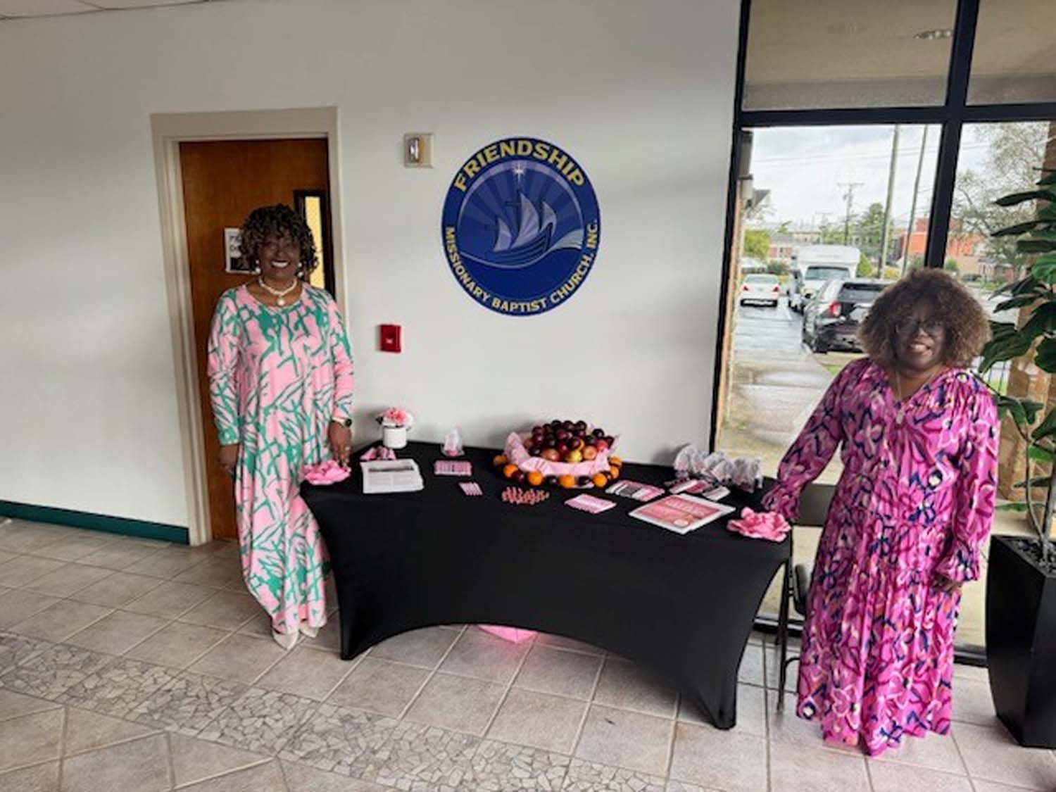 Two people in colorful patterned dresses stand on either side of a table with treats, beneath a church logo on a wall.