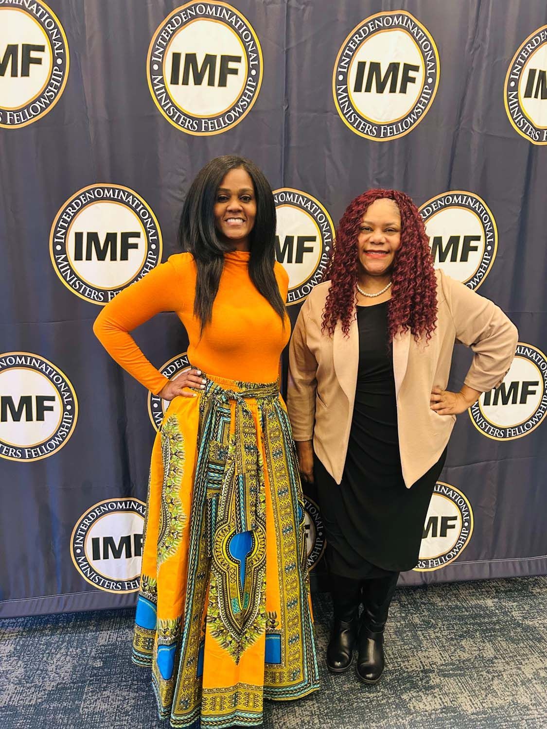 Two women stand smiling in front of a blue backdrop featuring the repeating International Monetary Fund (IMF) logo.