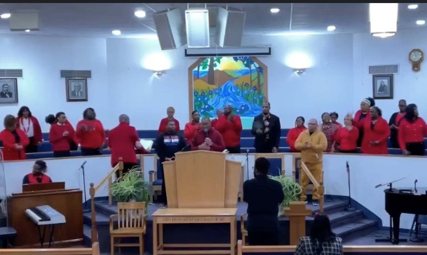 A choir in red shirts stands on a stage in front of a colorful mural in a church, with musicians and a leader present.
