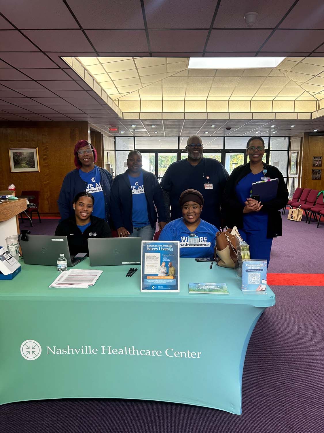 Six staff members from Nashville Healthcare Center stand behind a blue table in a large hall, smiling at the camera.