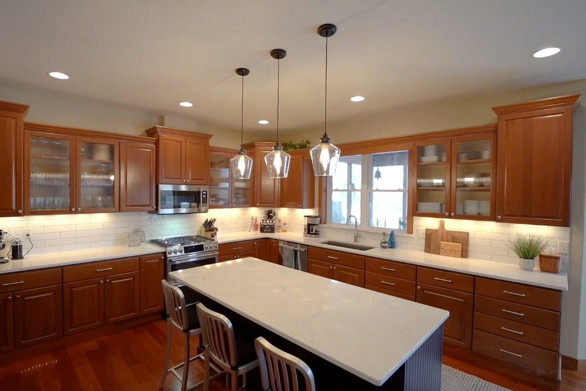 Warm wood kitchen with glass-front upper cabinets, white quartz countertops, dark island, subway tile backsplash, pendant lights, and stainless appliances.