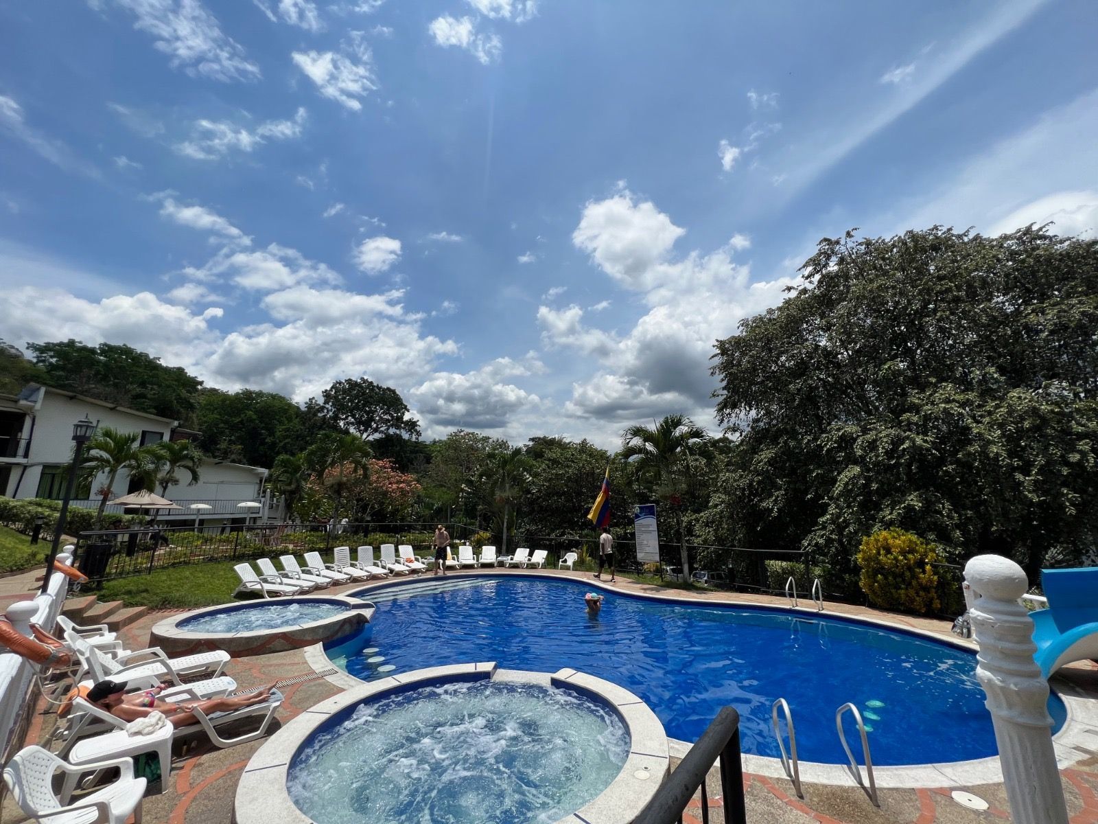 Zona de piscina con agua azul, tumbonas blancas y exuberantes árboles verdes bajo un cielo brillante.