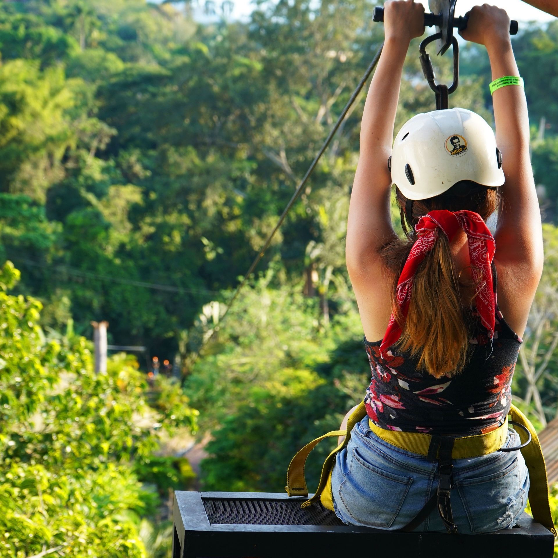Persona en tirolina, con los brazos en alto. Lleva casco y pañuelo rojo. Al fondo, exuberantes árboles verdes.