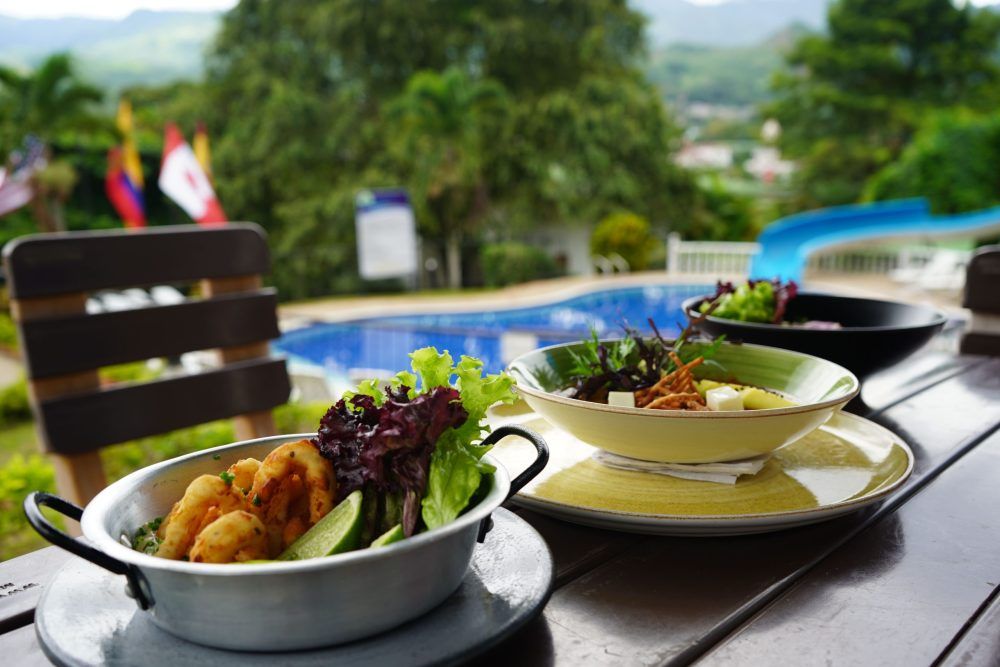 Platos de comida sobre una mesa de madera con vistas a una piscina y árboles, banderas al fondo.