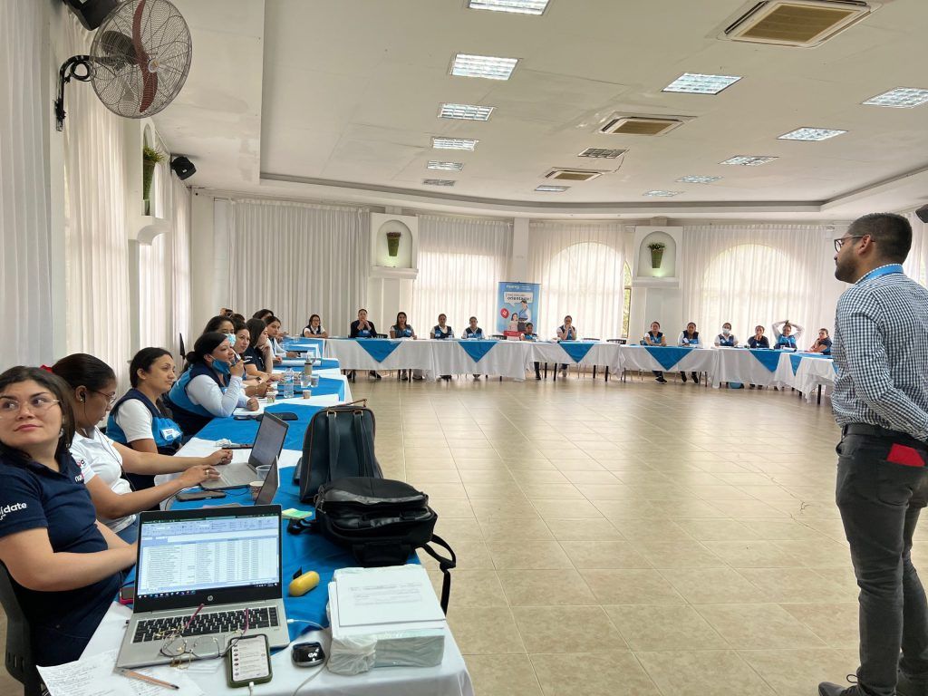 Reunión en una sala grande. Las personas están sentadas alrededor de una mesa larga; un hombre habla. Se ven ordenadores portátiles y papeles.