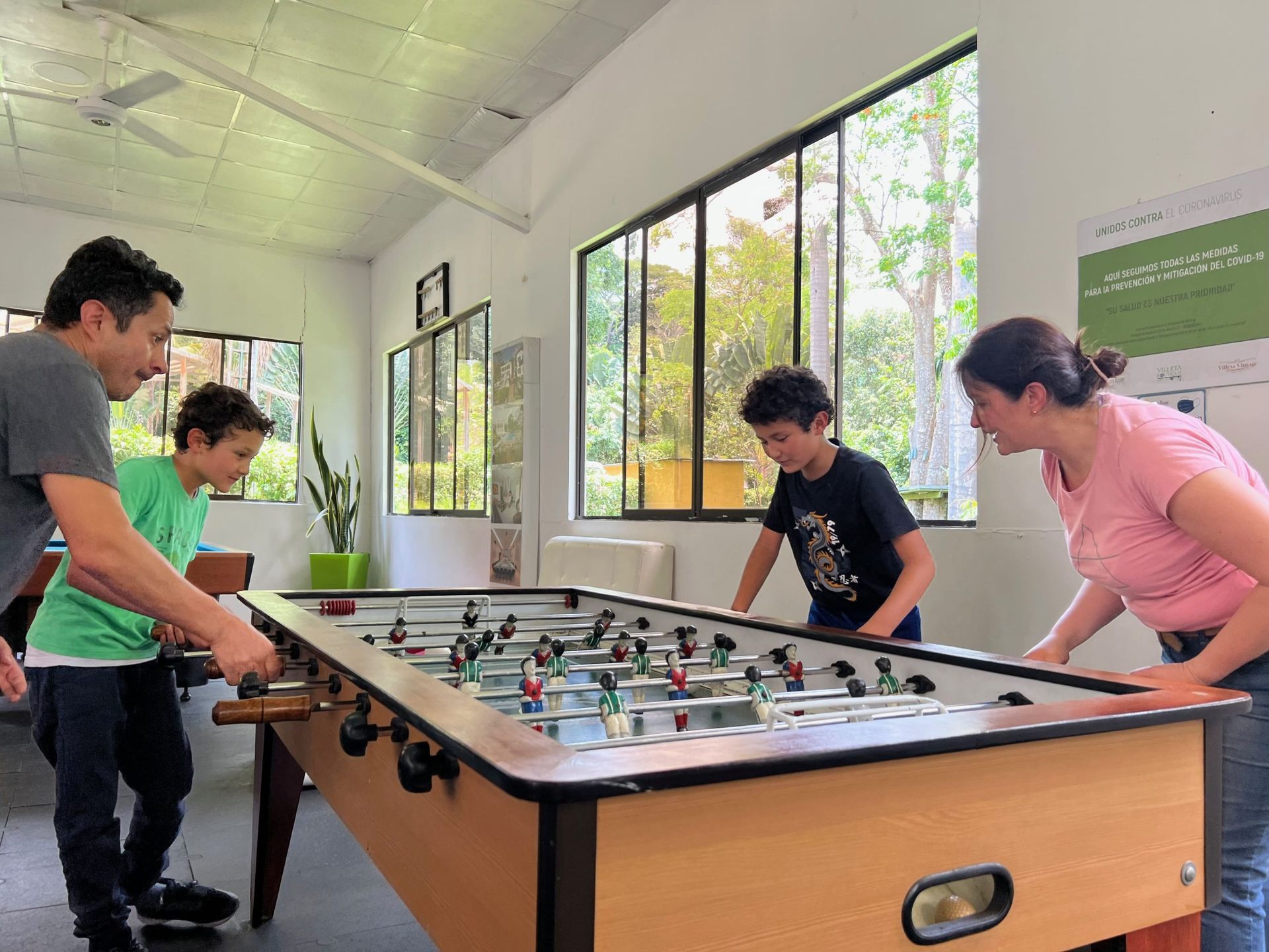 Una familia juega al futbolín en una habitación con ventanas. Dos niños y dos adultos, sonrientes, participan en el juego.
