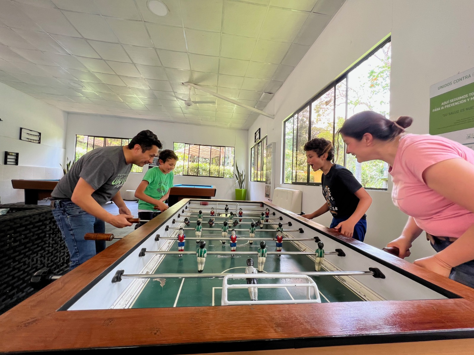 Una familia juega al futbolín en una habitación bien iluminada con ventanas. Parecen concentrados y sonrientes.