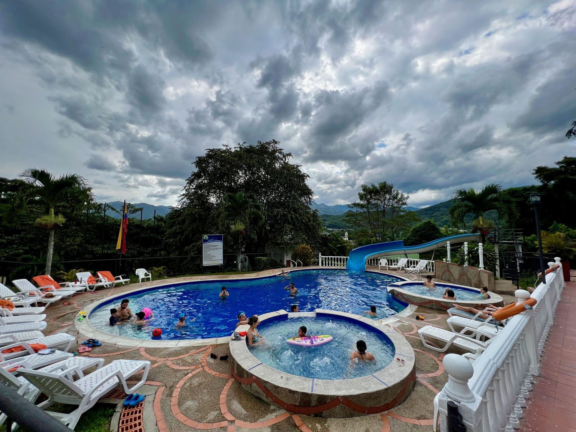 Piscina con gente bajo un cielo nublado. Se ven tumbonas, un tobogán y una montaña al fondo.