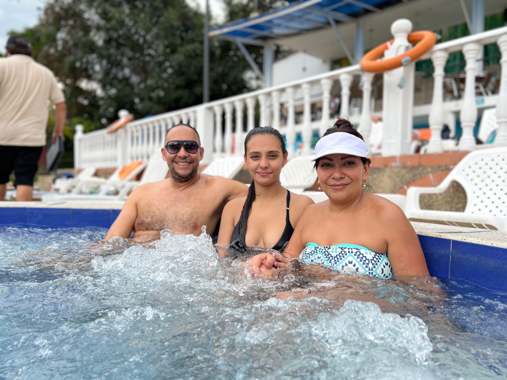 Tres personas sonriendo en un jacuzzi. Un hombre con gafas de sol. Una mujer en traje de baño. El lugar es una piscina.