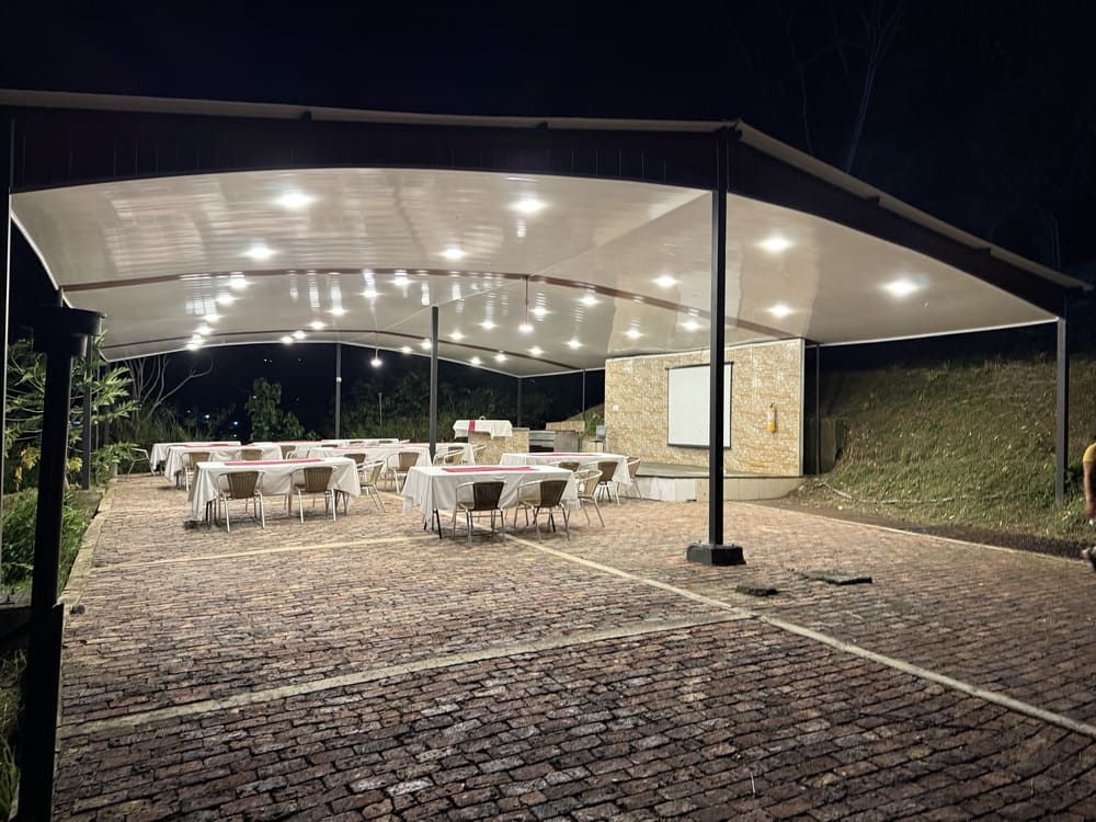 Comedor al aire libre bajo un toldo, con mesas preparadas para una comida; iluminado por luces superiores.
