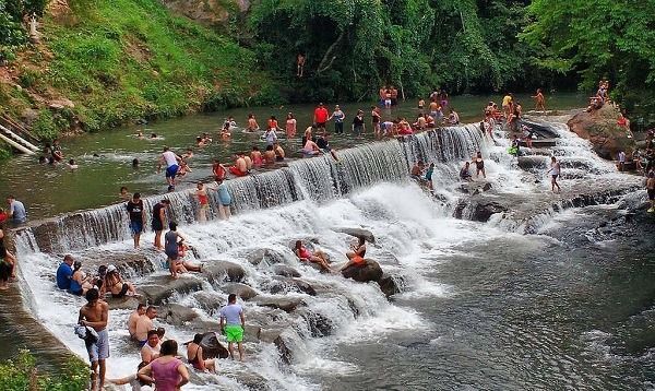 Personas nadando y sentadas en una cascada escalonada en un río, rodeadas de vegetación verde.