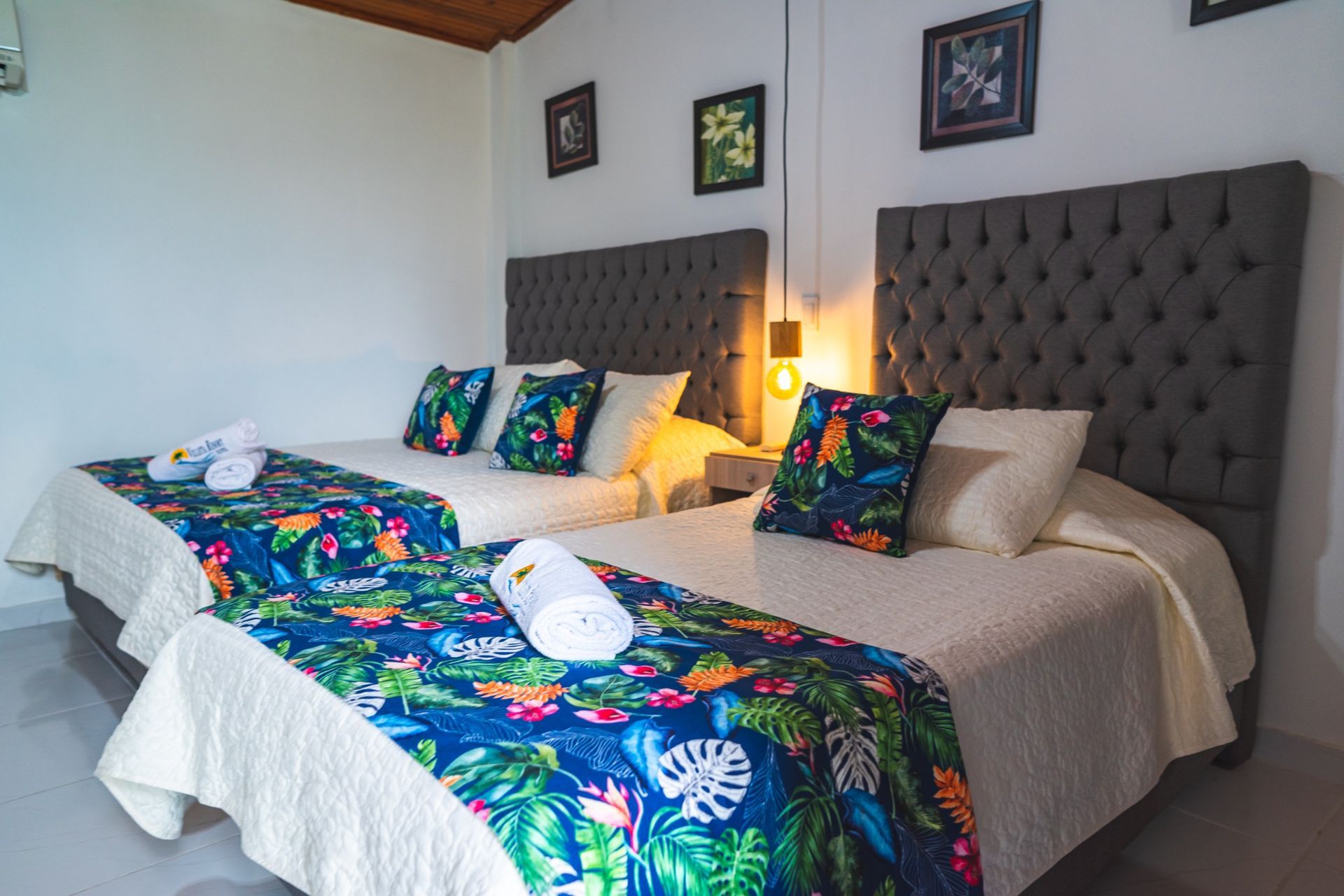 Dos camas con caminos de cama florales, cabeceros grises y sábanas blancas en una habitación luminosa.