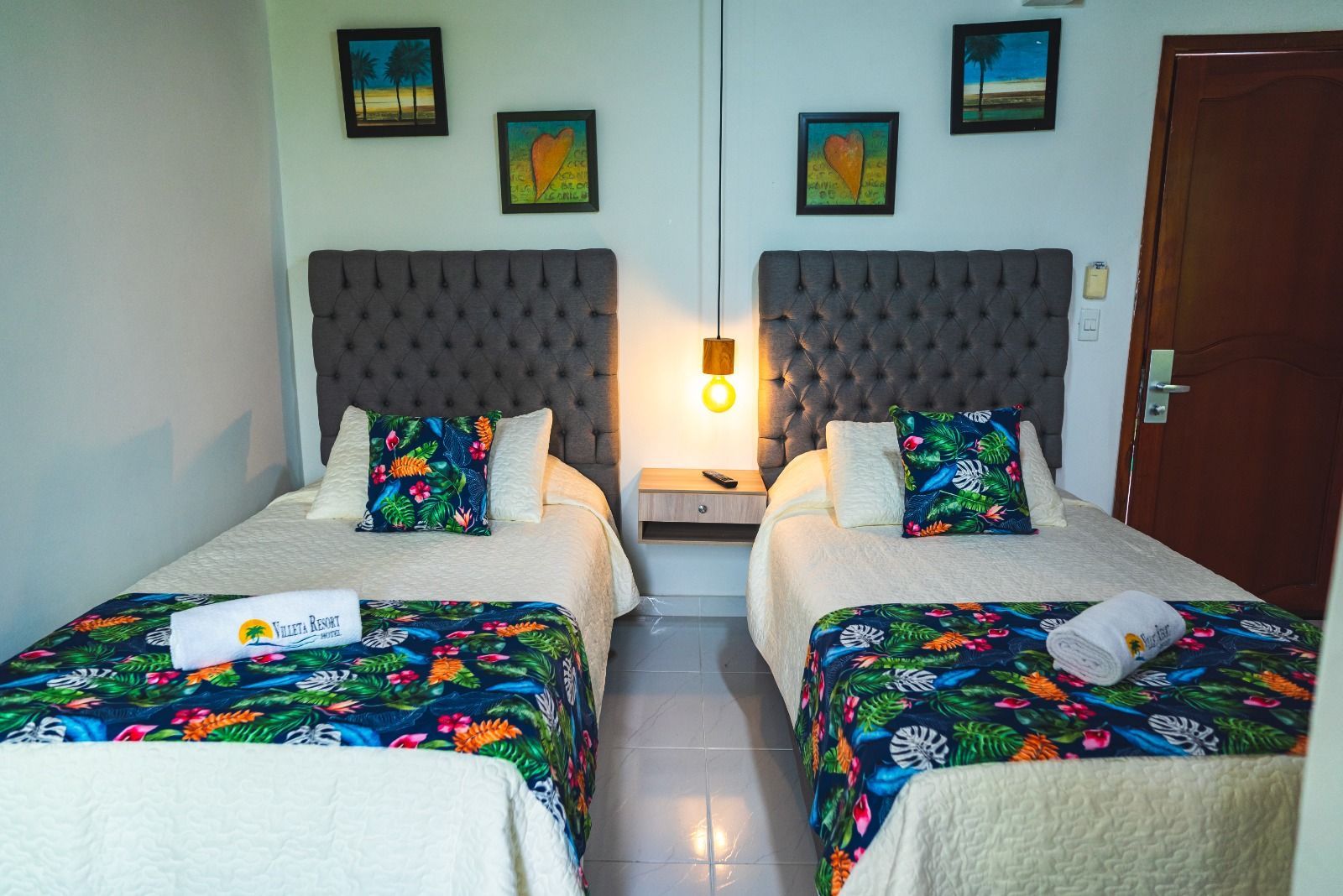 Dos camas individuales en una habitación con cabeceros capitoné grises, mesita de noche, colchas florales y obras de arte en la pared.