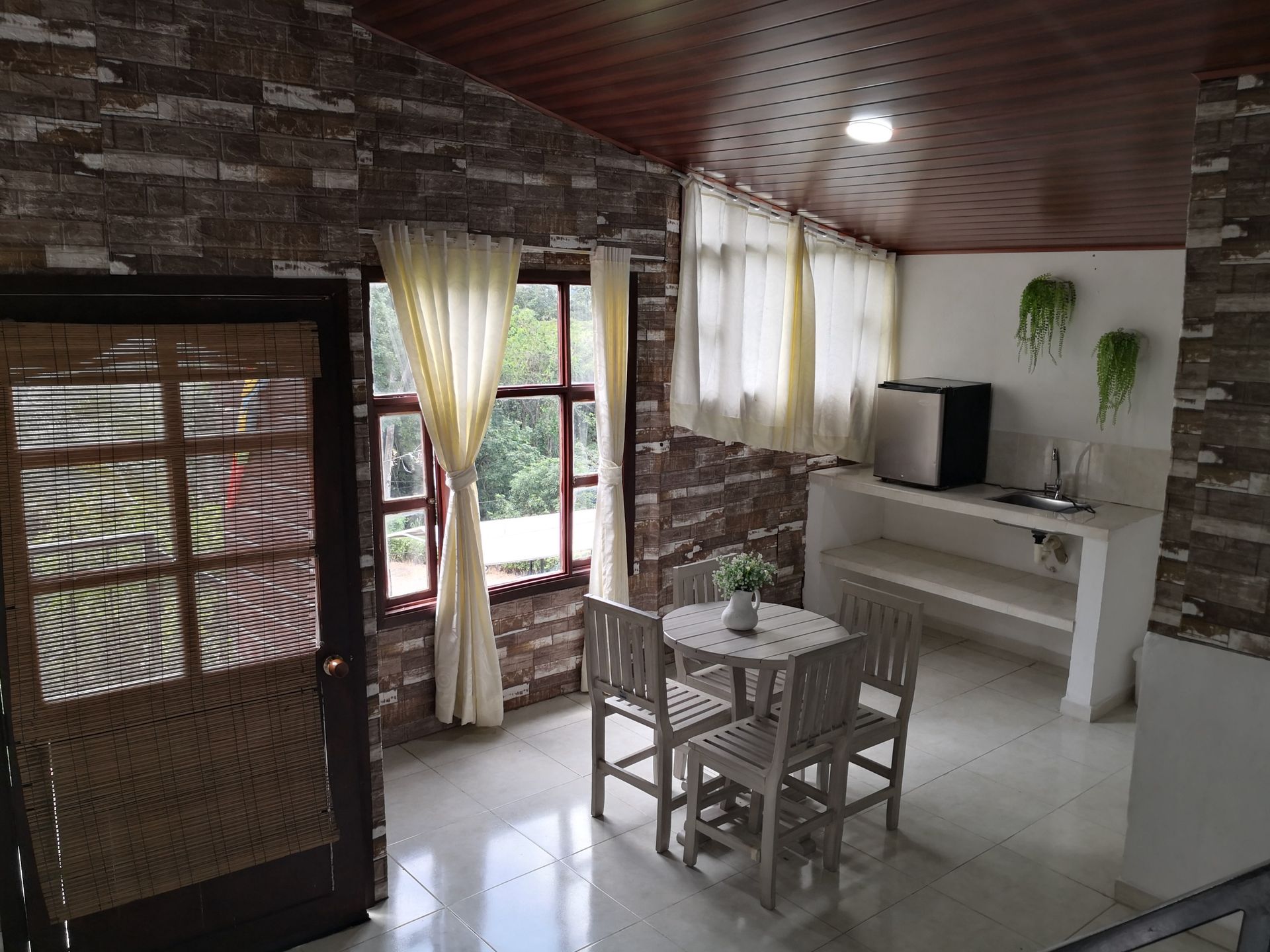 Interior de una habitación con paredes de piedra, ventanas, mesa y sillas, y un pequeño refrigerador.