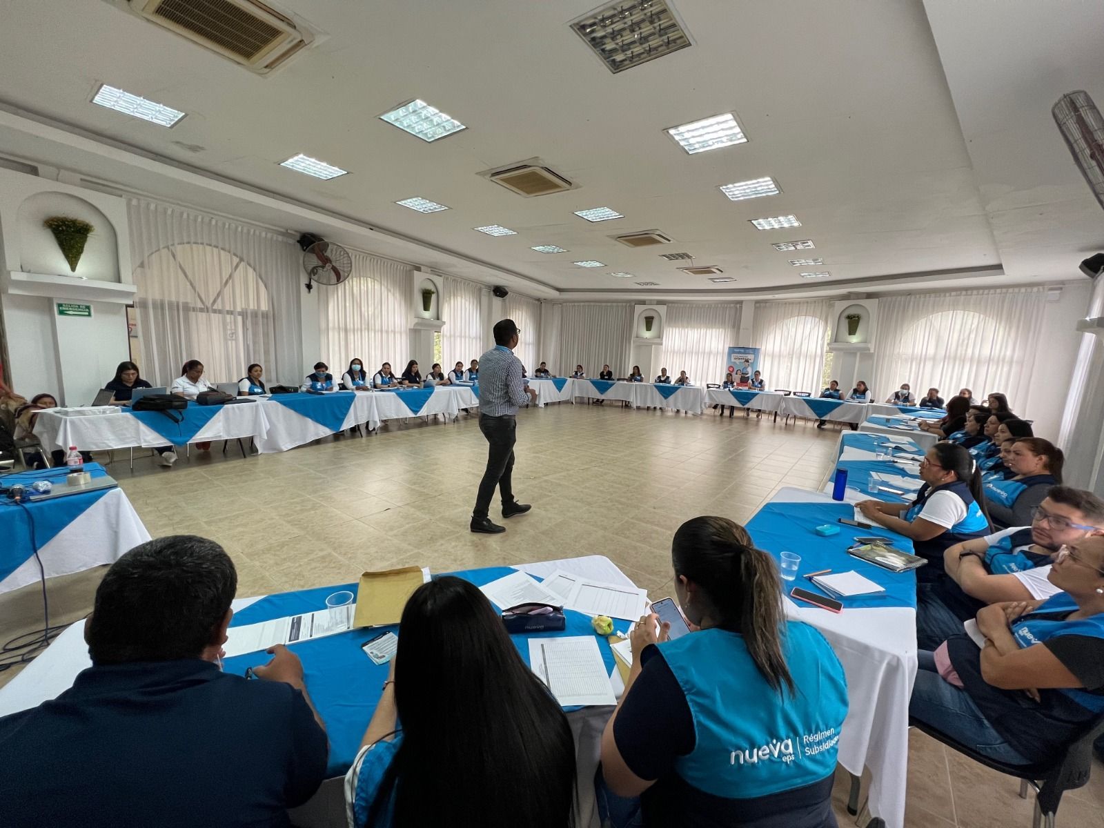 Hombre realizando una presentación ante un grupo en una mesa de conferencias, numerosos asistentes, colores azul y blanco, ambiente luminoso.