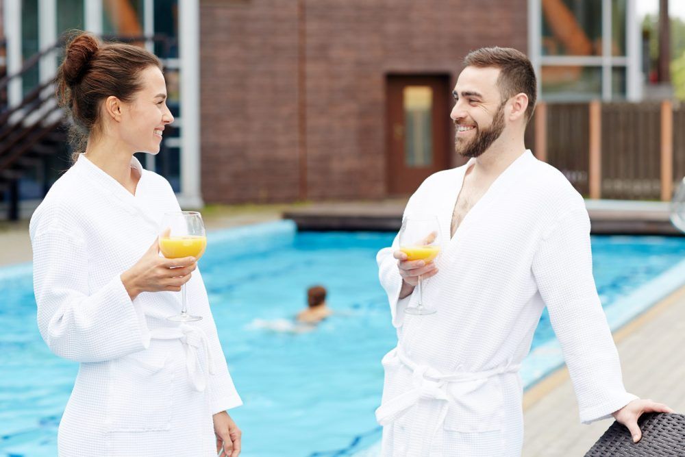 Pareja en batas blancas junto a una piscina, sonriendo y con bebidas en la mano.