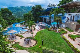 Vista aérea panorámica del complejo turístico con piscina, tobogán acuático y restaurante con vistas a una ladera boscosa.