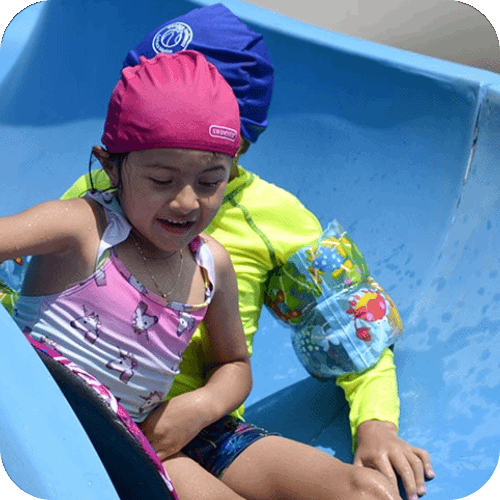 Una chica en traje de baño baja por un tobogán acuático azul junto a otra persona vestida de azul y amarillo.