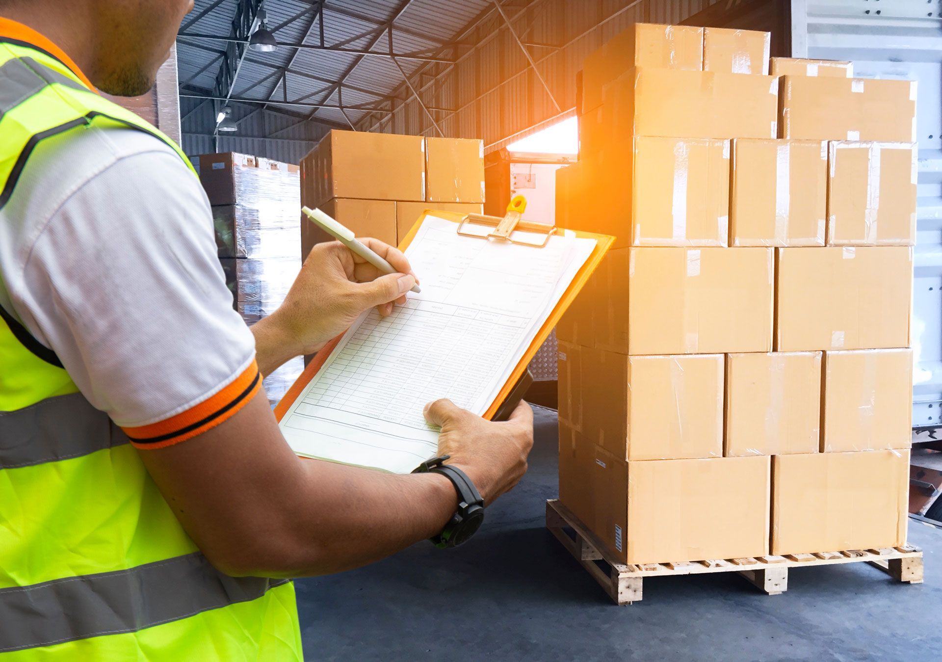 Warehouse worker in safety vest using clipboard to inspect boxes stacked on a pallet.