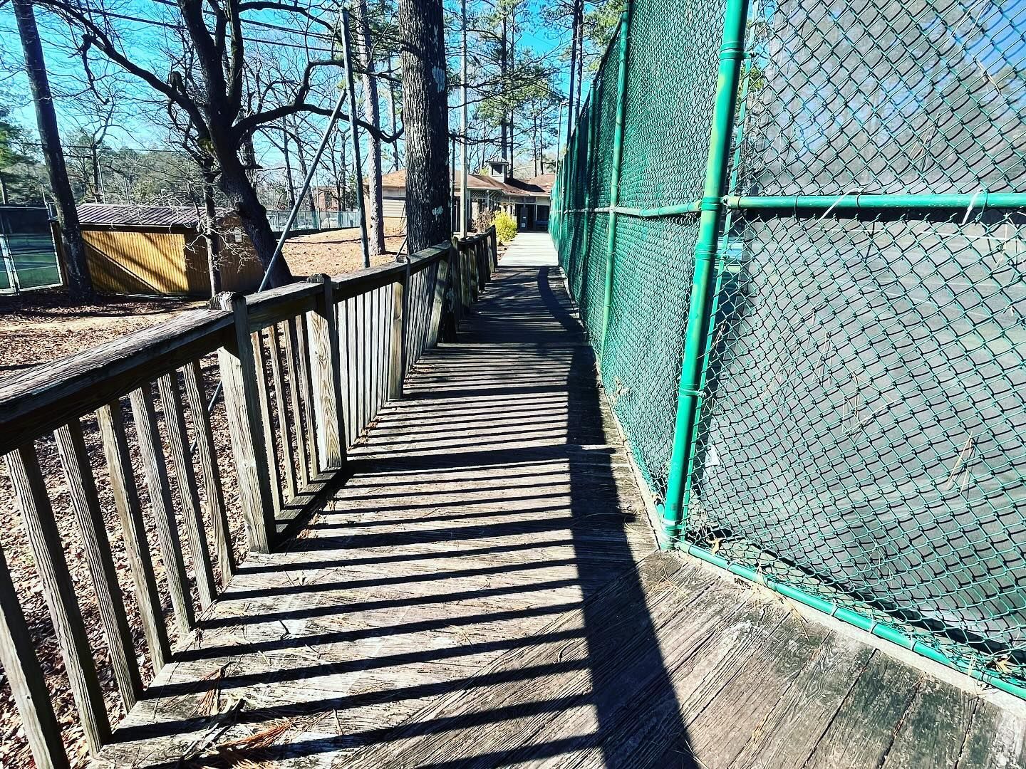 A wooden walkway leading to a tennis court behind a chain link fence.