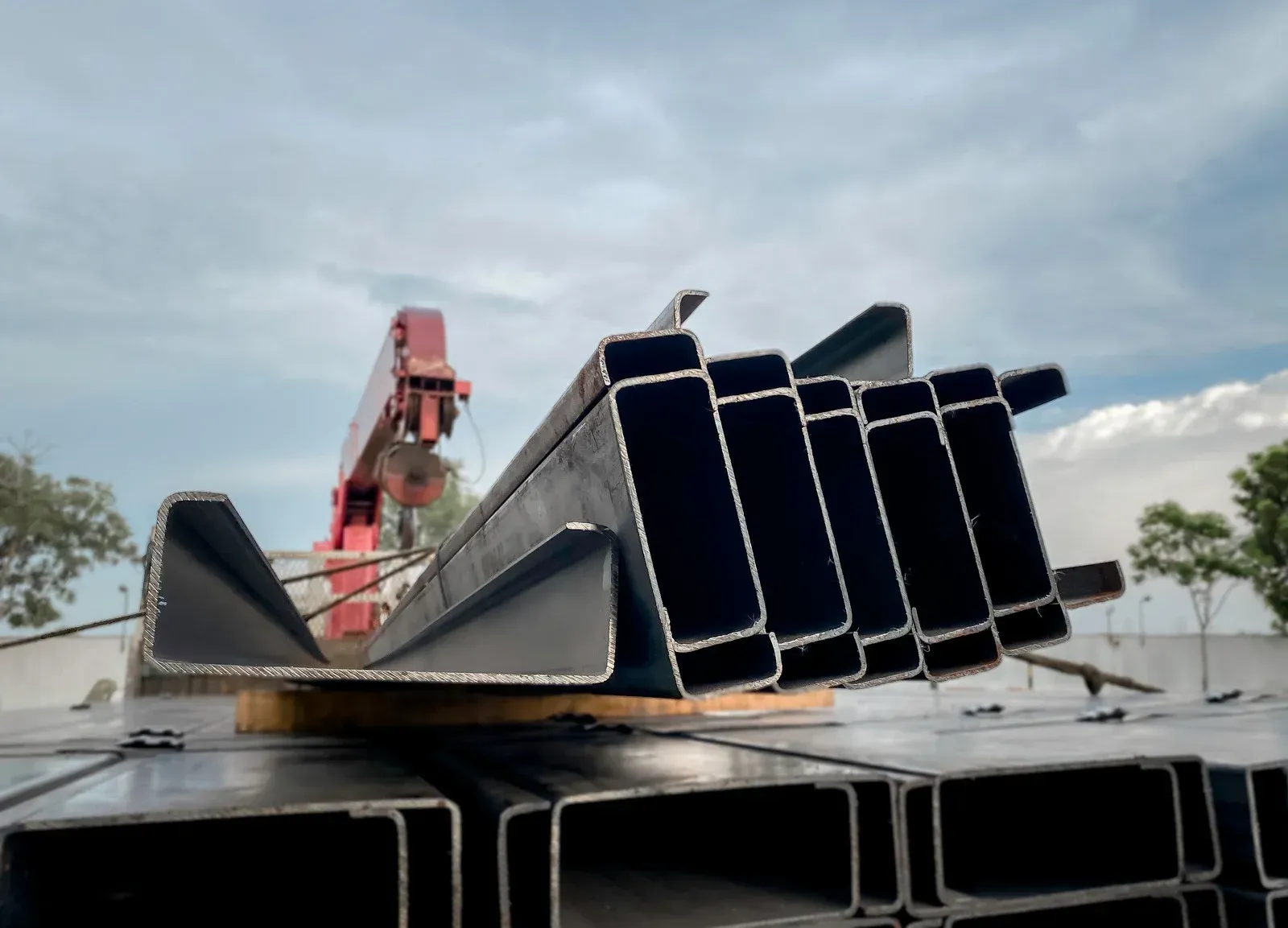 Steel C-beams stacked on a flatbed truck being loaded by a crane under a cloudy sky.
