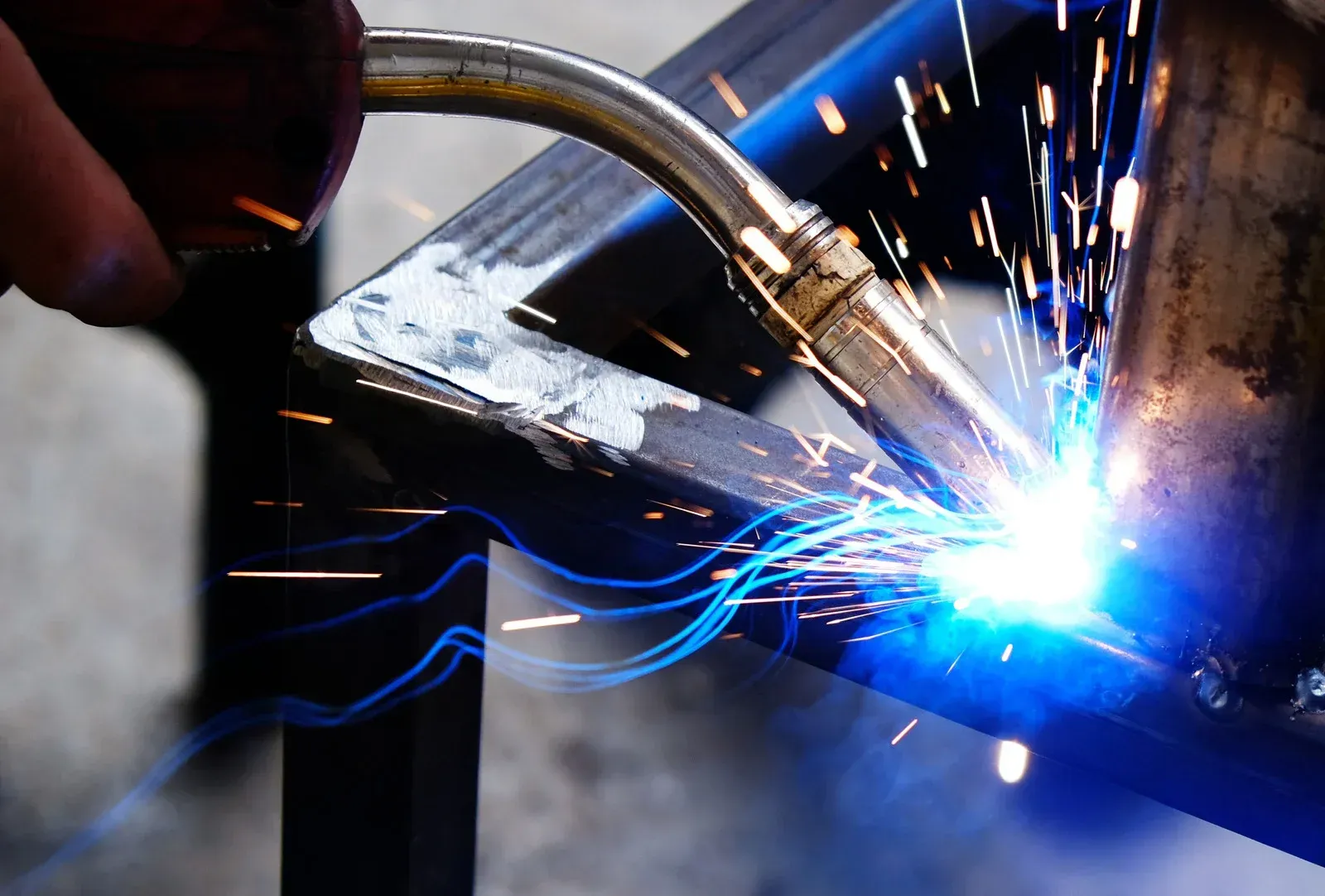 A welding torch joins two metal pieces, emitting a bright blue light and flying sparks against a dark background.