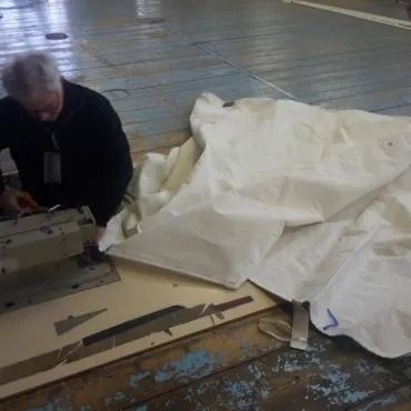 Uomo che lavora su una tela bianca, con attrezzi e ritagli su un tavolo. Il pavimento è in legno con vernice blu scrostata.