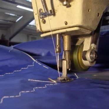 Primo piano di una macchina da cucire che cuce un tessuto blu con un motivo a zigzag, probabilmente in un laboratorio.