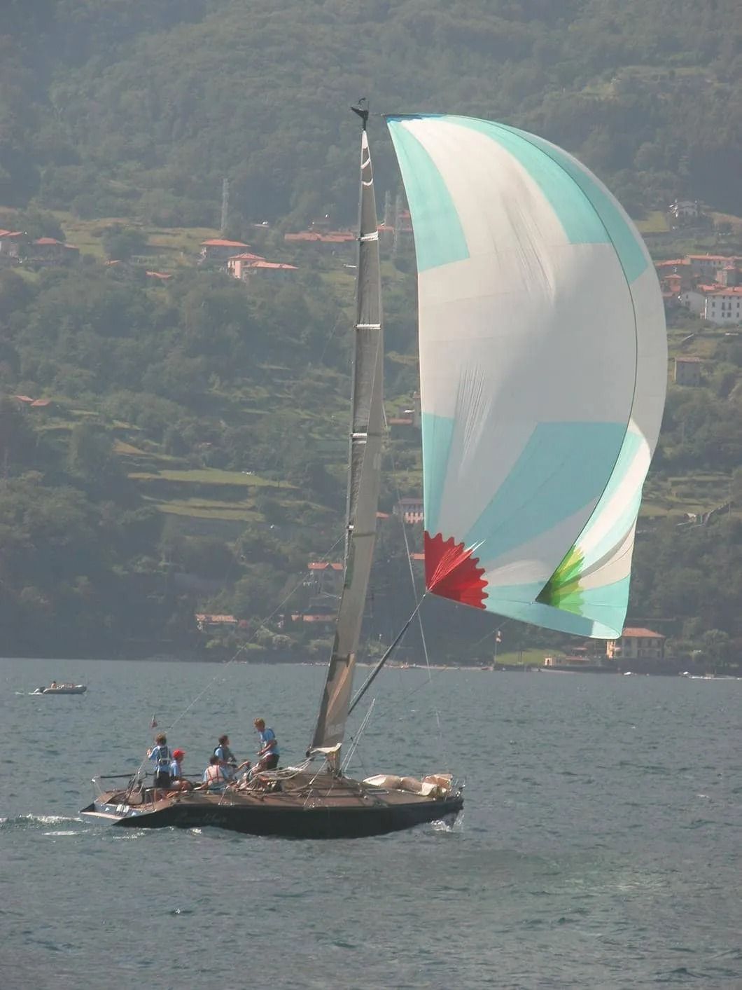 Barca a vela con spinnaker colorato in acqua, equipaggio visibile. Montagne e villaggio sullo sfondo.