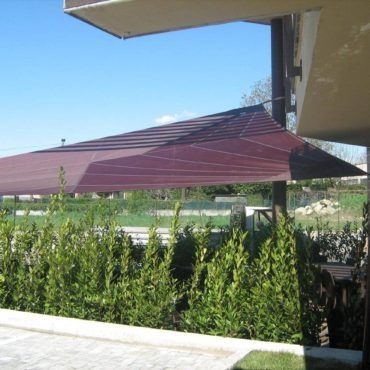 Una tenda da sole color bordeaux, fissata a un edificio, che fornisce ombra a un giardino con cespugli verdi.