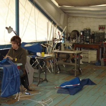 Una persona cuce un tessuto blu in un laboratorio. Una macchina da cucire, attrezzi e materiali riempiono lo spazio.