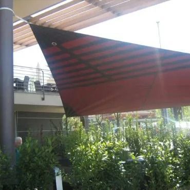 Vela ombreggiante triangolare rossa che crea ombra su un patio. Travi nere e colonna marrone.