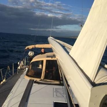 Barca a vela in mare con una persona al timone. La vela è parzialmente spiegata, con nuvole scure in cielo.