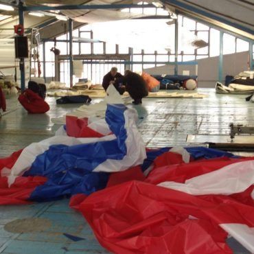 Paracadute con i colori rosso, bianco e blu steso sul pavimento di un ambiente interno; persone sullo sfondo.