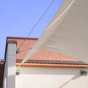 Vela parasole color crema fissata all'angolo di un edificio, con un muro di mattoni e un cielo azzurro sullo sfondo.