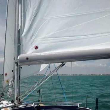 Una barca a vela con vele bianche su un mare blu. Il boma è teso e in lontananza si vede una costa.