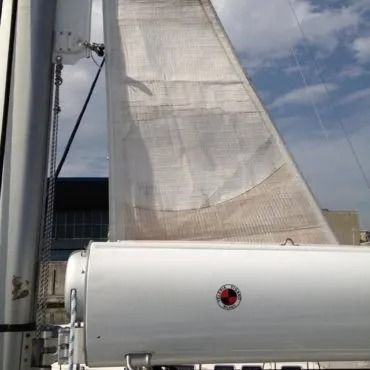 La vela bianca di una barca a vela è spiegata, accanto a un oggetto cilindrico bianco, sullo sfondo di un cielo nuvoloso.