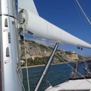 Albero e boma di una barca a vela con il Mar Mediterraneo e le scogliere costiere sullo sfondo.