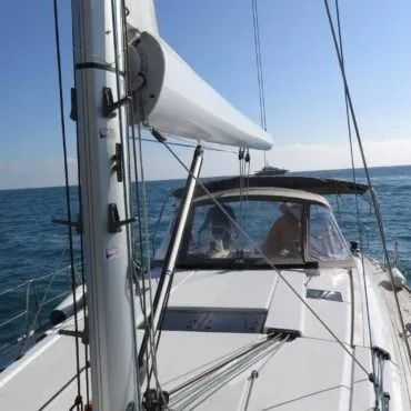 Barca a vela in acqua con albero e vela issati. Una persona in pozzetto timona la barca in una giornata di sole.